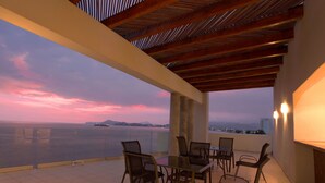 Terraço com mesa e cadeiras, com vista para um corpo d'água e terra distante ao pôr do sol.