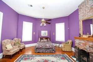 A room with purple walls, a bed with floral bedding, two armchairs, a fireplace, and a ceiling fan. - Esplanade Suites (New Orleans)