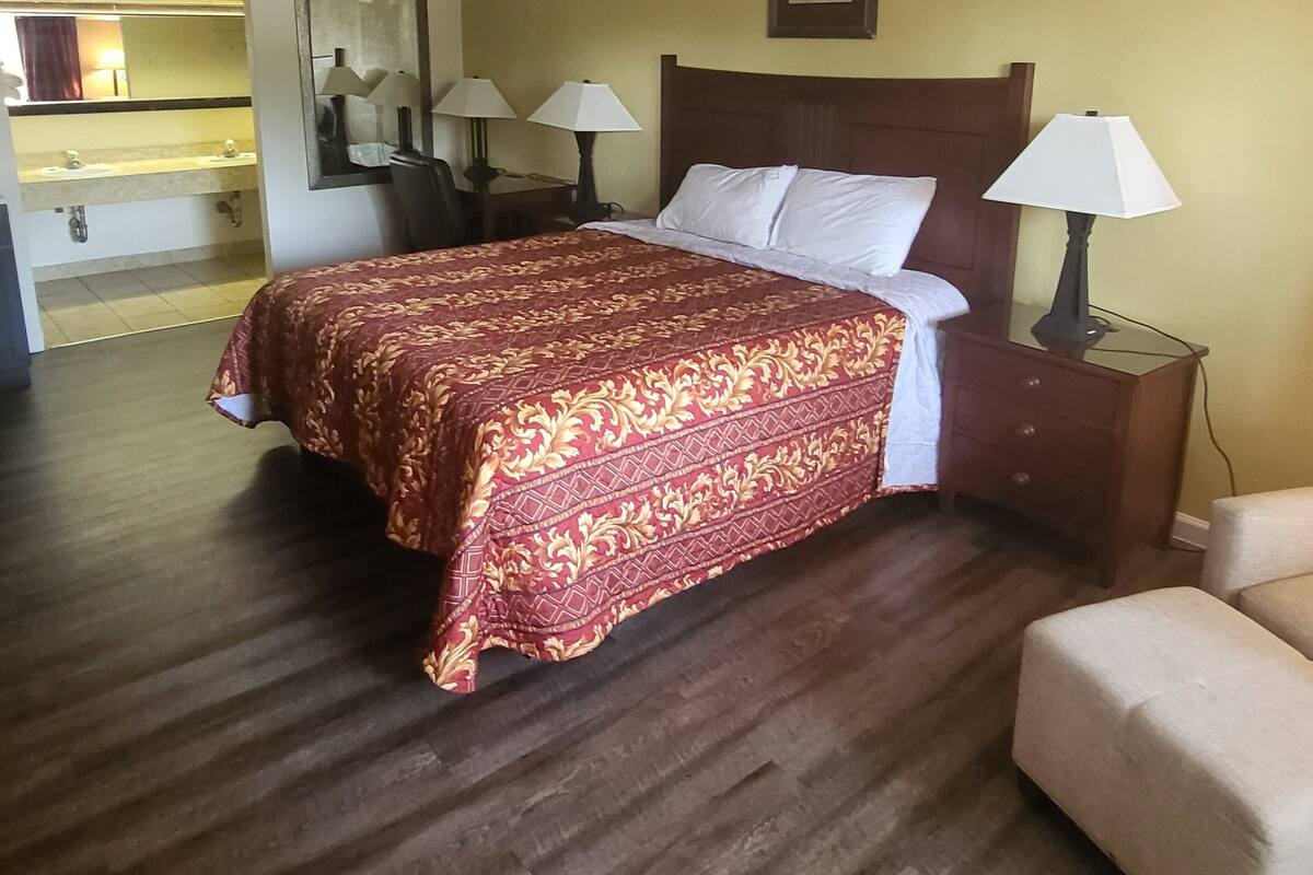 A hotel room with a bed, two bedside tables with lamps, a chair, a mirror, and a bathroom visible through an open door.