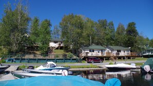 Una cabaña junto al lago con muelle, barcos y un coche estacionado cerca.