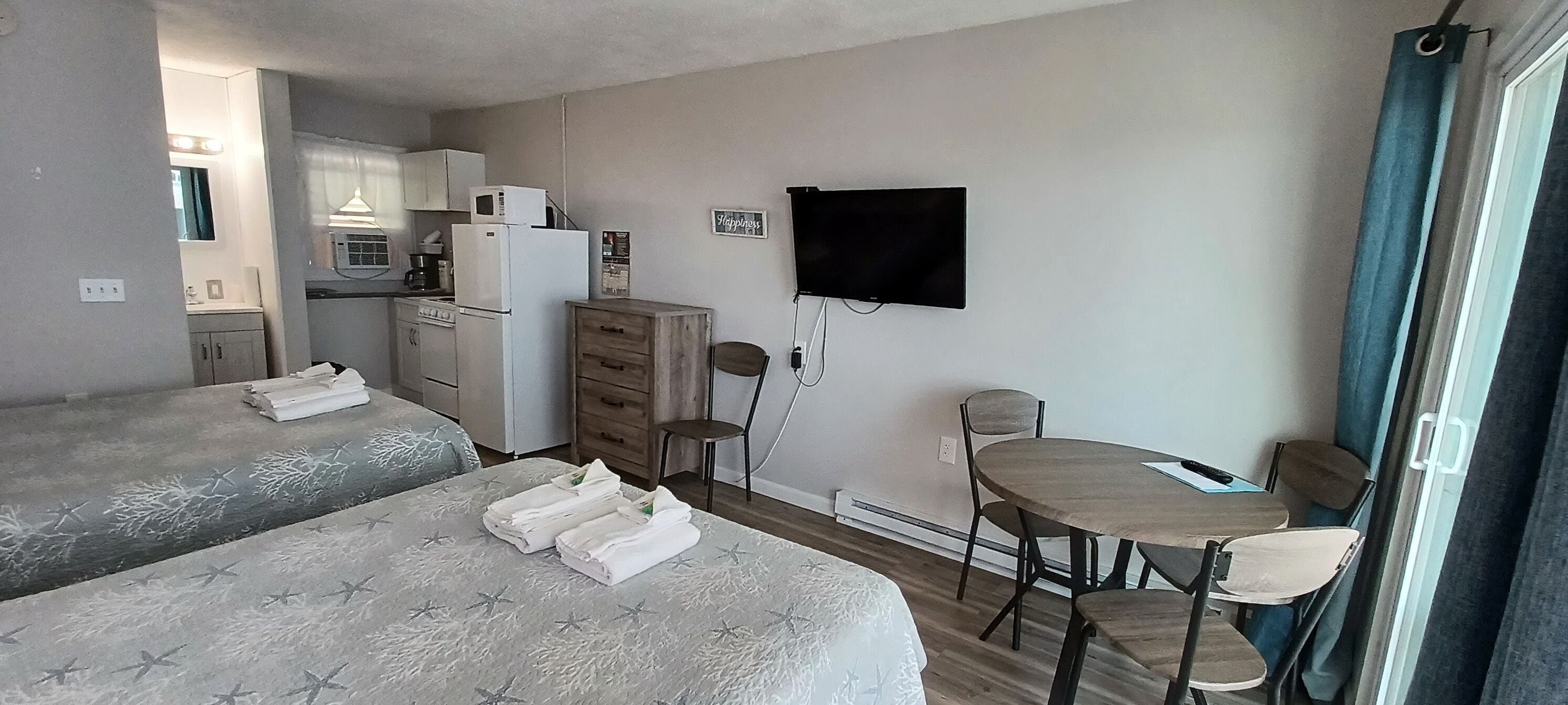A compact hotel room with two beds, a small kitchenette, a TV, and a dining area.