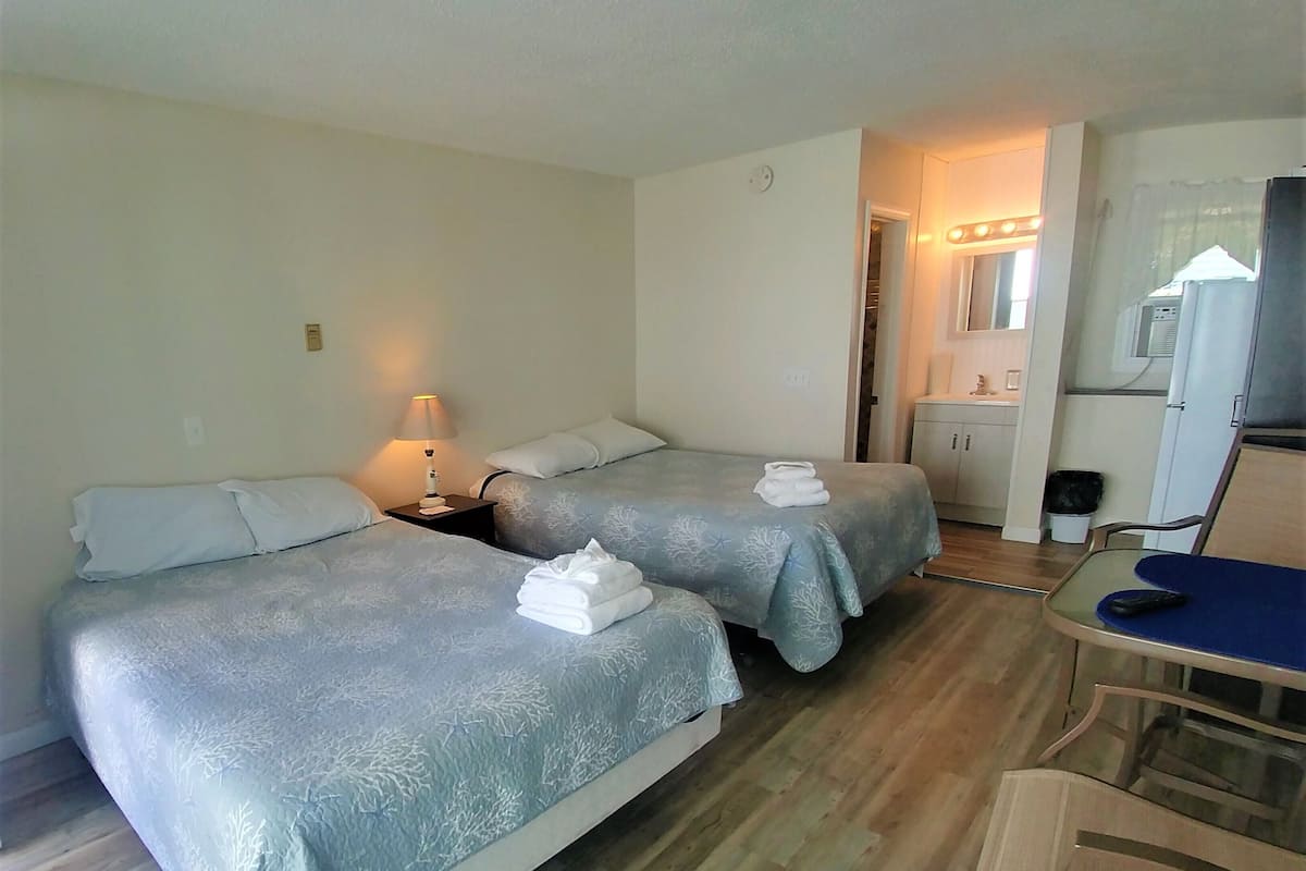 Ein Hotelzimmer mit zwei Betten, einem Schreibtisch, einem Stuhl, einer kleinen Küchenzeile und einem durchgegangene Tür sichtbaren Badezimmer.