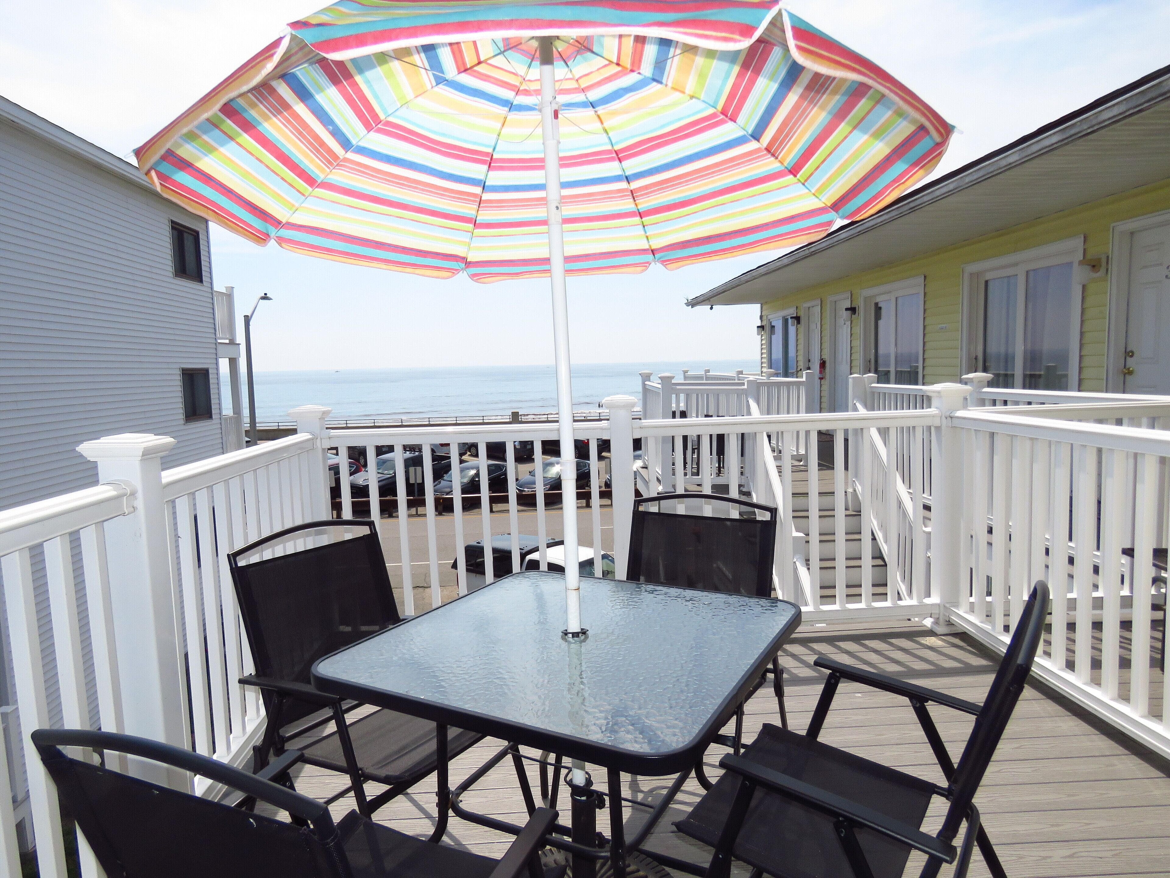 A deck with a glass table and chairs, a colorful striped umbrella, and a view of the ocean.