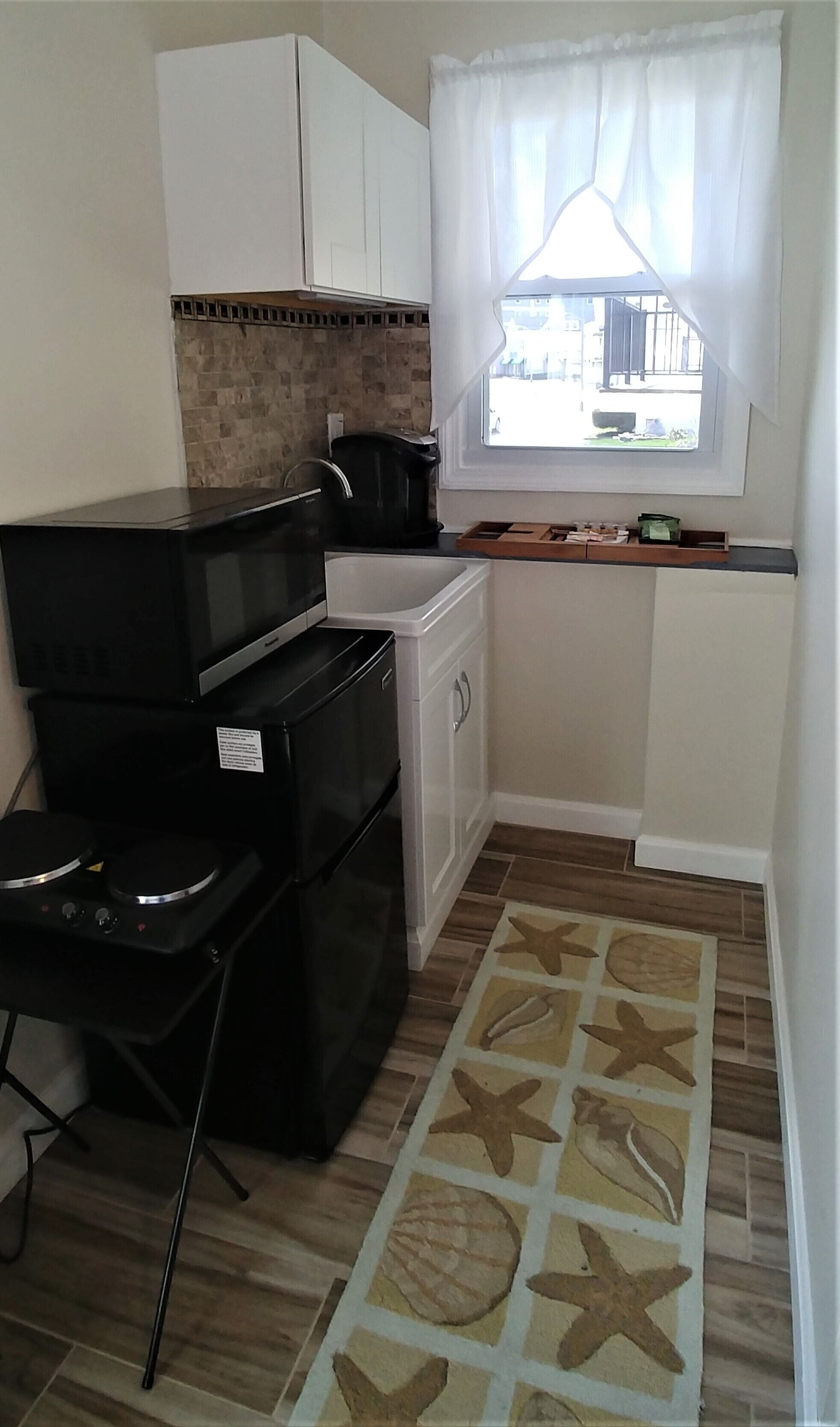 A compact kitchen with a microwave, stove, and small refrigerator. There's a window with curtains, a tiled backsplash, and a wooden countertop.
