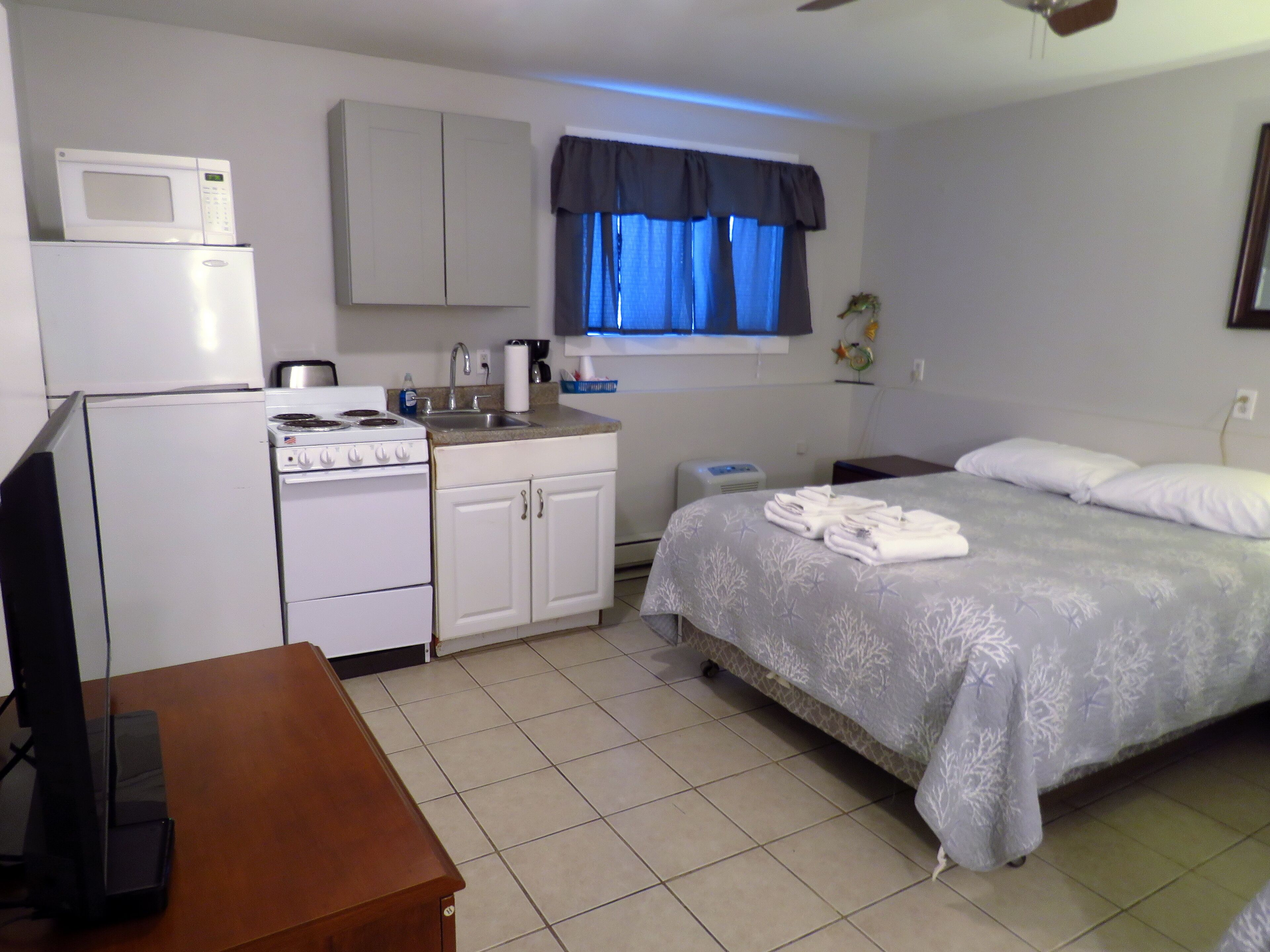 A compact hotel room with a kitchenette, a bed with a grey floral bedspread, a wooden desk, and a ceiling fan.
