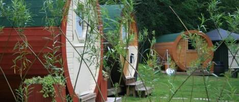 A row of rustic, cylindrical wooden cabins with a green roof, surrounded by a grassy area with bamboo plants.