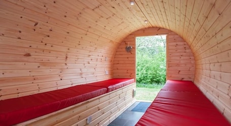 A wooden cabin with a curved ceiling, a bench with a red cushion, and a door leading to an outdoor area.