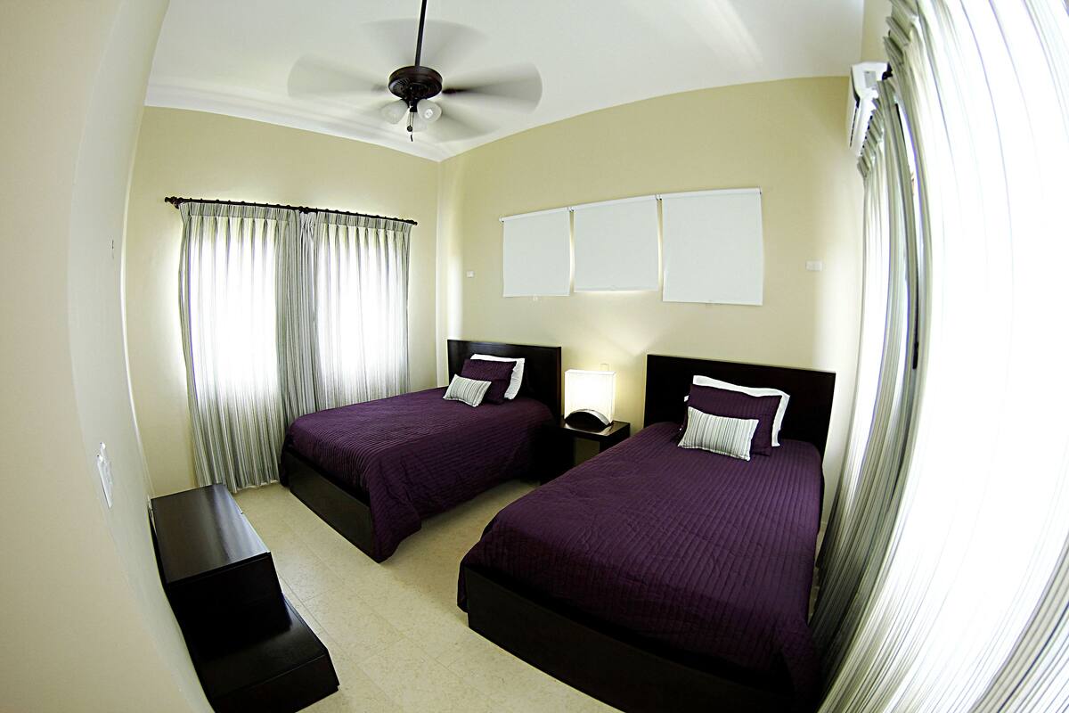 A hotel room with two beds, a ceiling fan, a nightstand with a lamp, and a window with curtains.