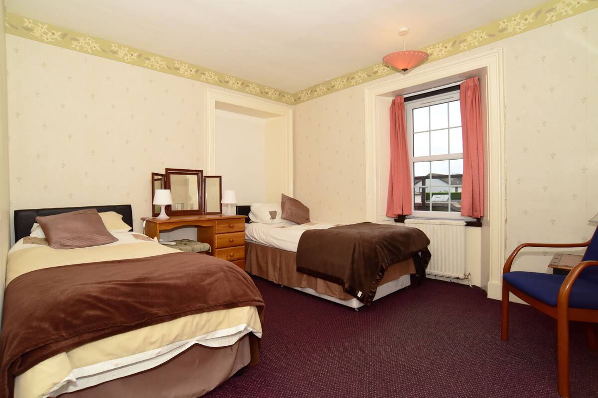A hotel room with two beds, a desk, a chair, a window with curtains, and a radiator.