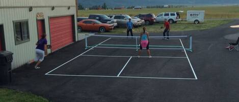 A tennis court with a net, surrounded by parked cars and a building with a red garage door.