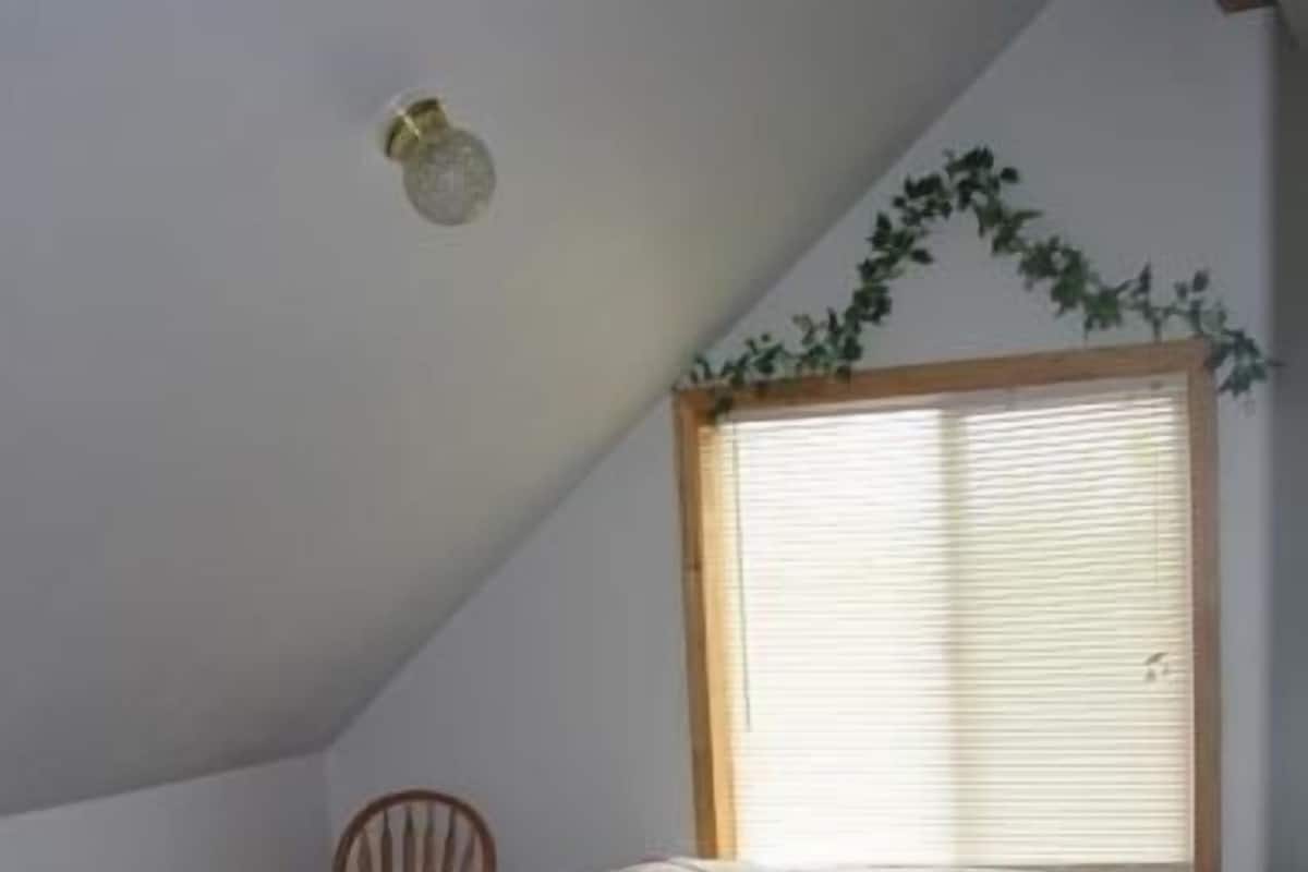A bedroom with a sloped ceiling, a single bed with floral bedding, a chair, a window with blinds, and a small plant on the wall.