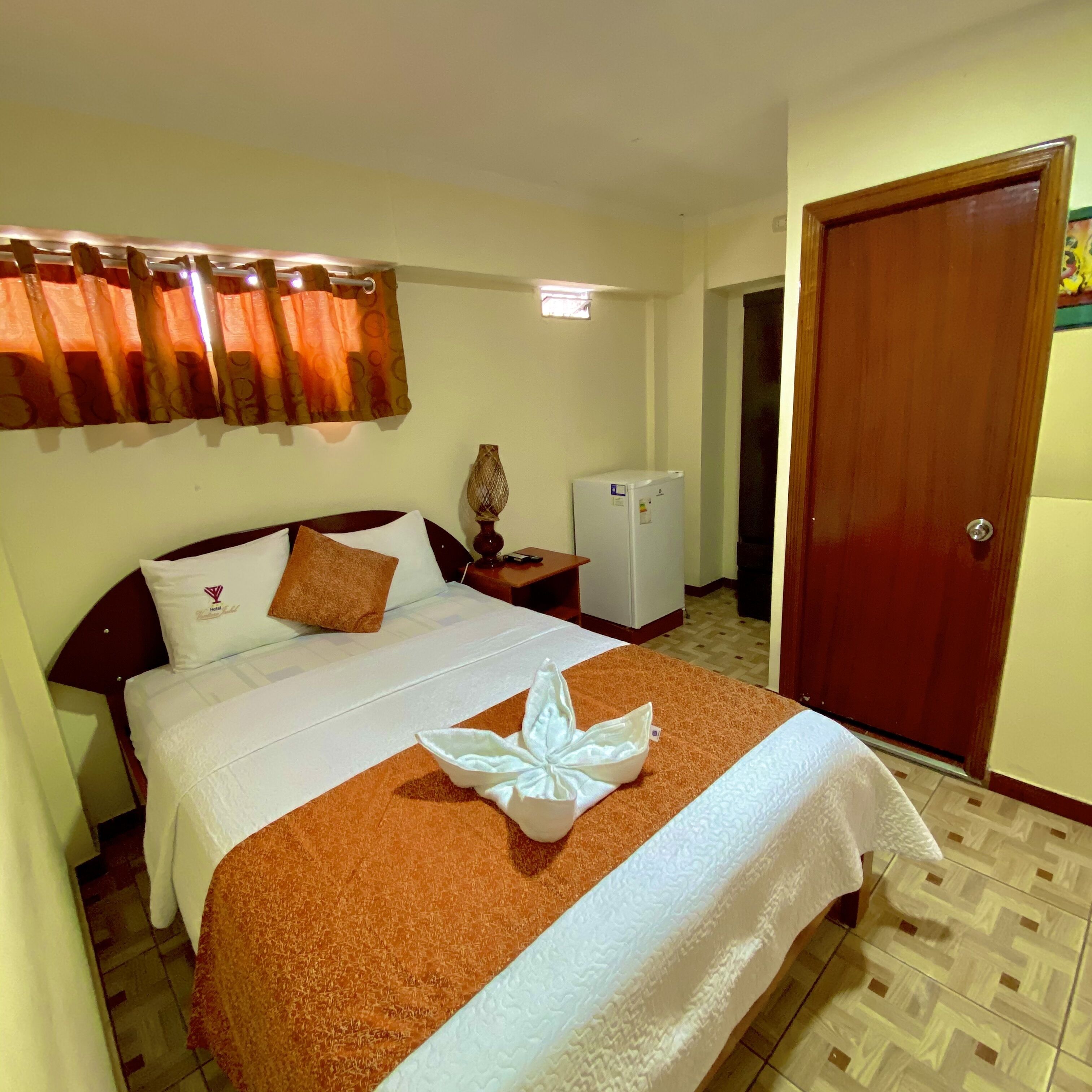 A hotel room with a bed, a nightstand, a small refrigerator, and a door.