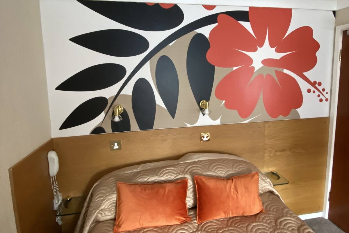 A hotel room with a bed, two pillows, a wooden headboard, a wall with a floral design, and a telephone on the side.