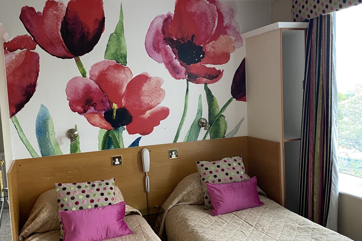A hotel room with two beds, each with a quilted beige bedspread and a pillow with a polka dot pattern. A large floral mural adorns the wall behind the beds.