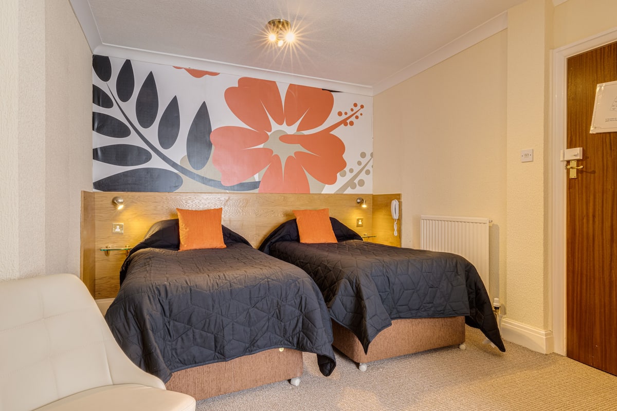 A hotel room with two beds, a wooden headboard, a floral wall mural, a white chair, and a wooden door.