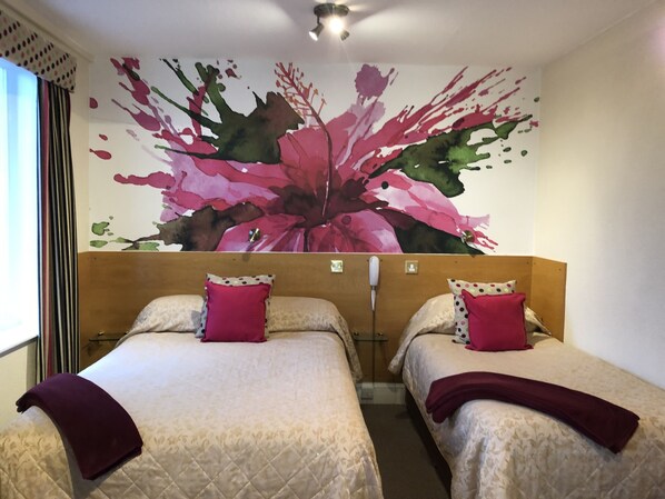 A hotel room with two beds, each with a headboard and a red pillow. The room features a large floral artwork on the wall above the beds. - Park Manor Hotel (Scarborough)