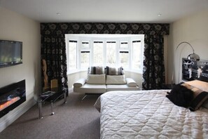A modern hotel room with a large bed, a seating area with a sofa and pillows, a fireplace, and a large window with curtains.