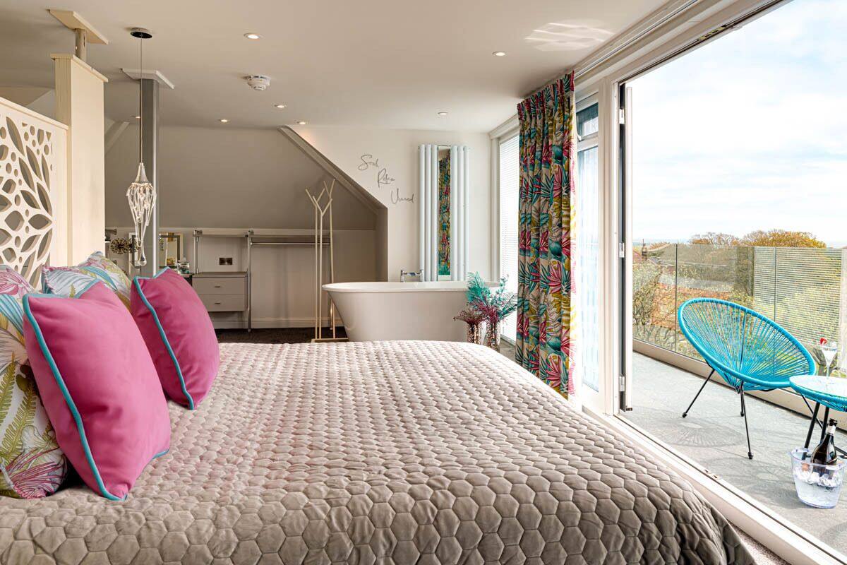 A bedroom with a large bed, a bathroom, and a balcony with a chair and table.