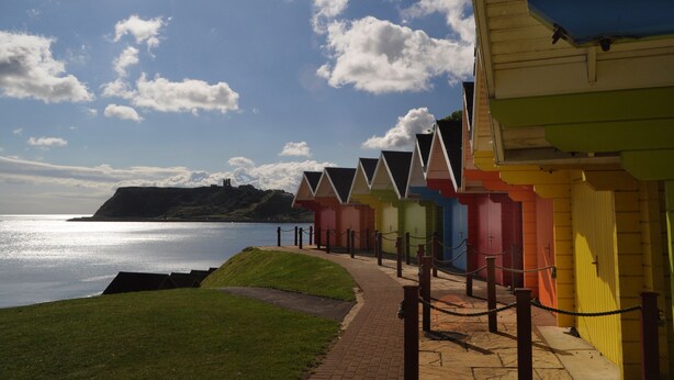 A row of colorful beach huts along a seaside path with a view of the ocean and hills in the background. - Park Manor Hotel (Scarborough)