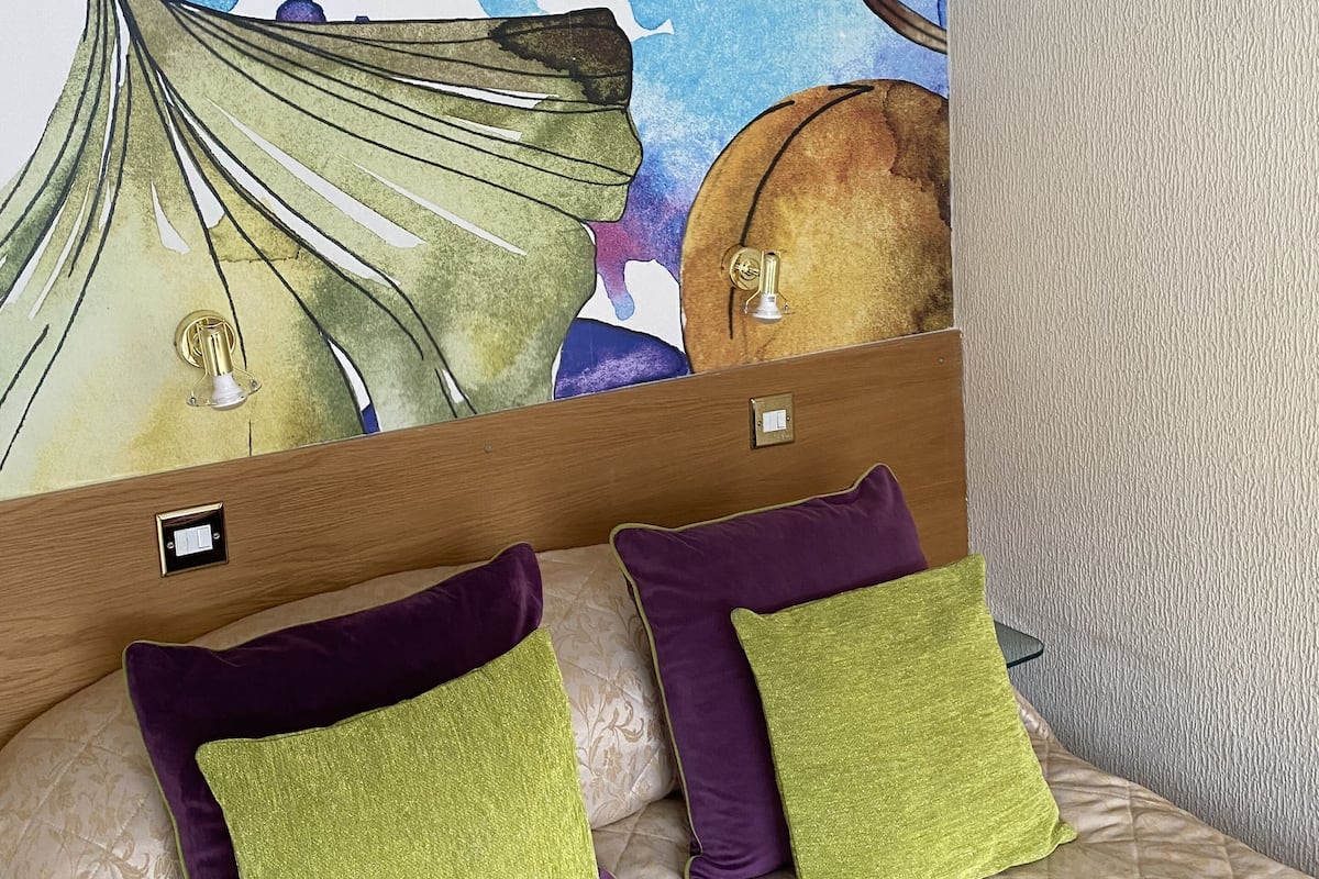 A bedroom with a large wall mural, a wooden headboard, and two green and purple pillows on a beige bed.
