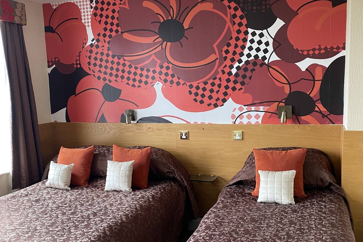 A hotel room with two beds, each with brown bedding and white pillows. A large floral-patterned wall mural is above the beds.