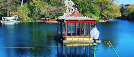 A small, enclosed pavilion with a roof, situated on a body of water, surrounded by trees and a distant building.