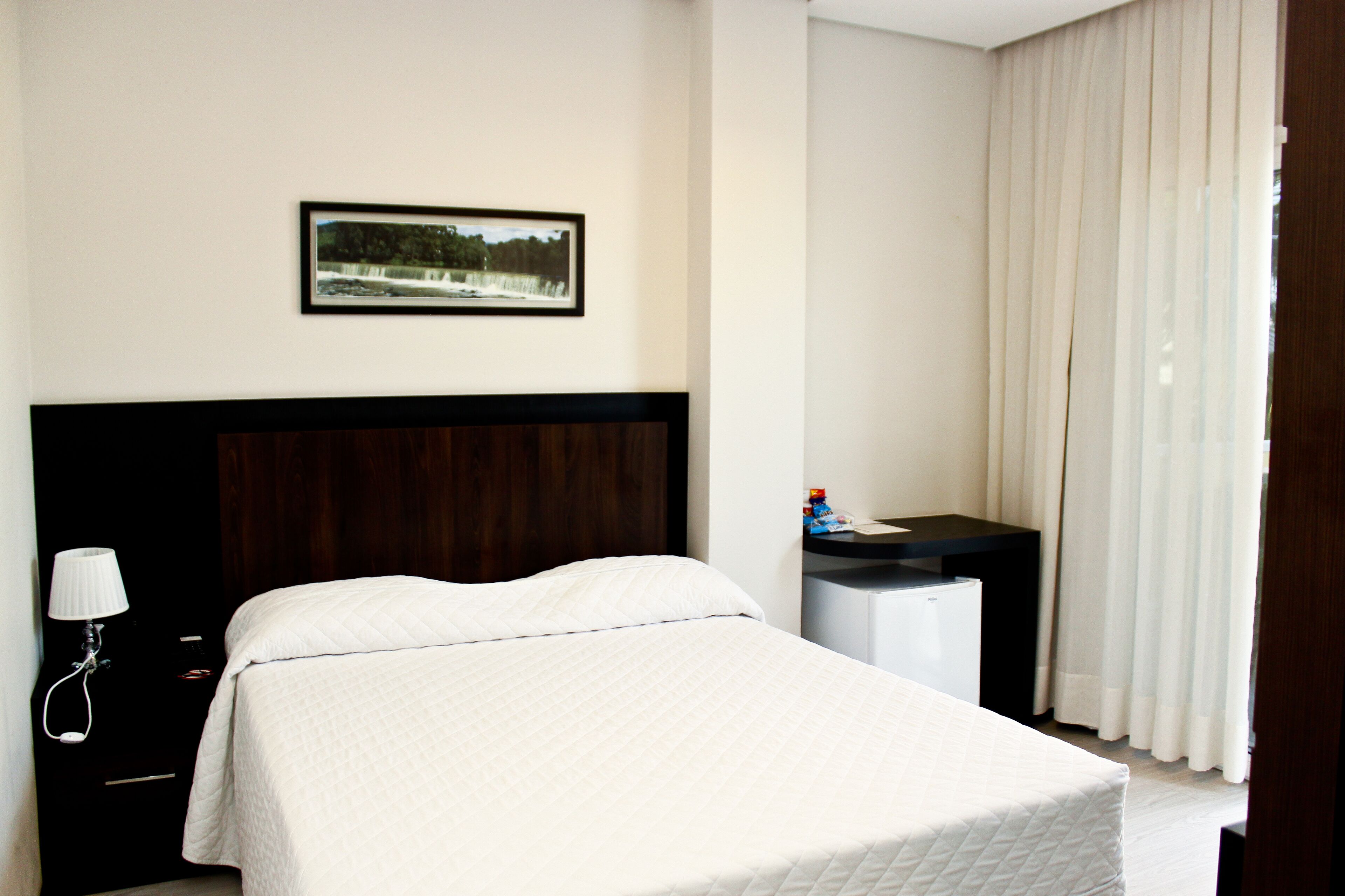 A hotel room with a bed, a desk, a lamp, a picture on the wall, and a window with curtains.