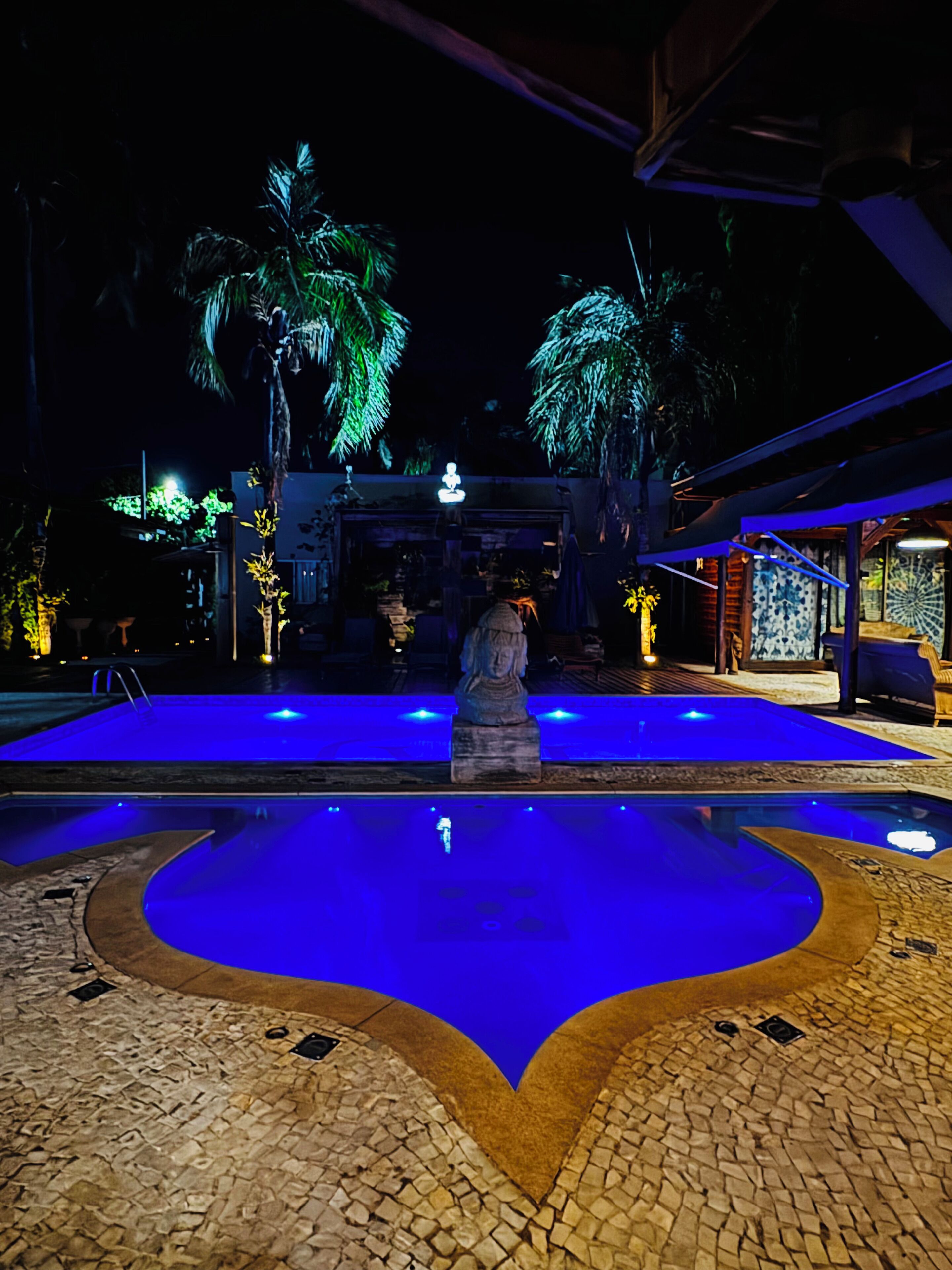 Un área de piscina nocturna con una estatua central, rodeada de palmeras y un suelo de baldosas de piedra.