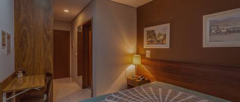 Quarto de hotel com uma escrivaninha de madeira, uma cadeira, uma cama com colcha estampada, um criado-mudo com abajur e duas fotos emolduradas na parede.