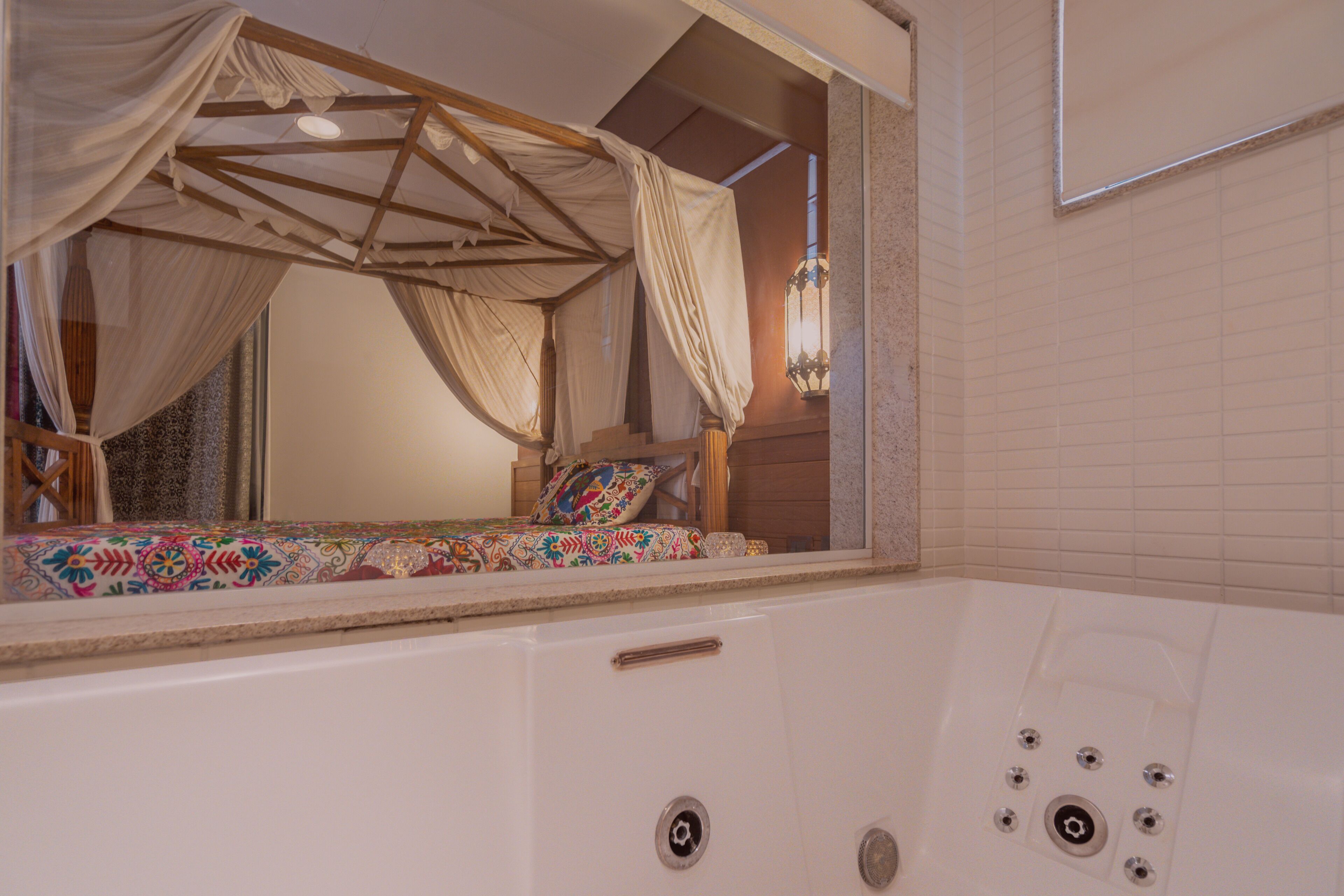 A room with a large window, a wooden canopy bed, a bathtub, and a mirror reflecting the bed.