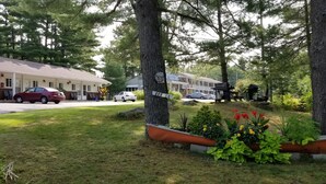 A hotel with a parking area, a red car, and a white car parked in front. There are trees and a flower bed with various plants. - Pinedale Inn (Gravenhurst)
