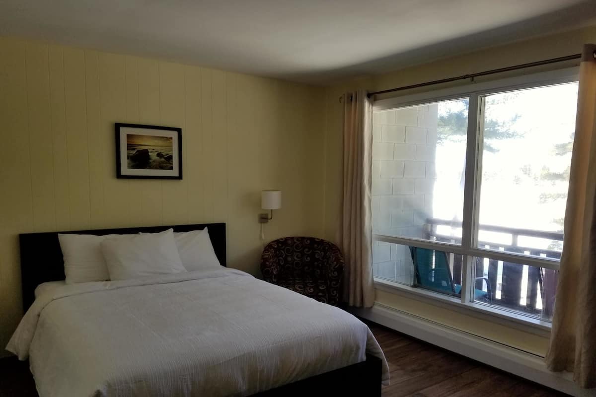 A hotel room with a bed, a chair, a window with a view of the outside, and a wall-mounted lamp.