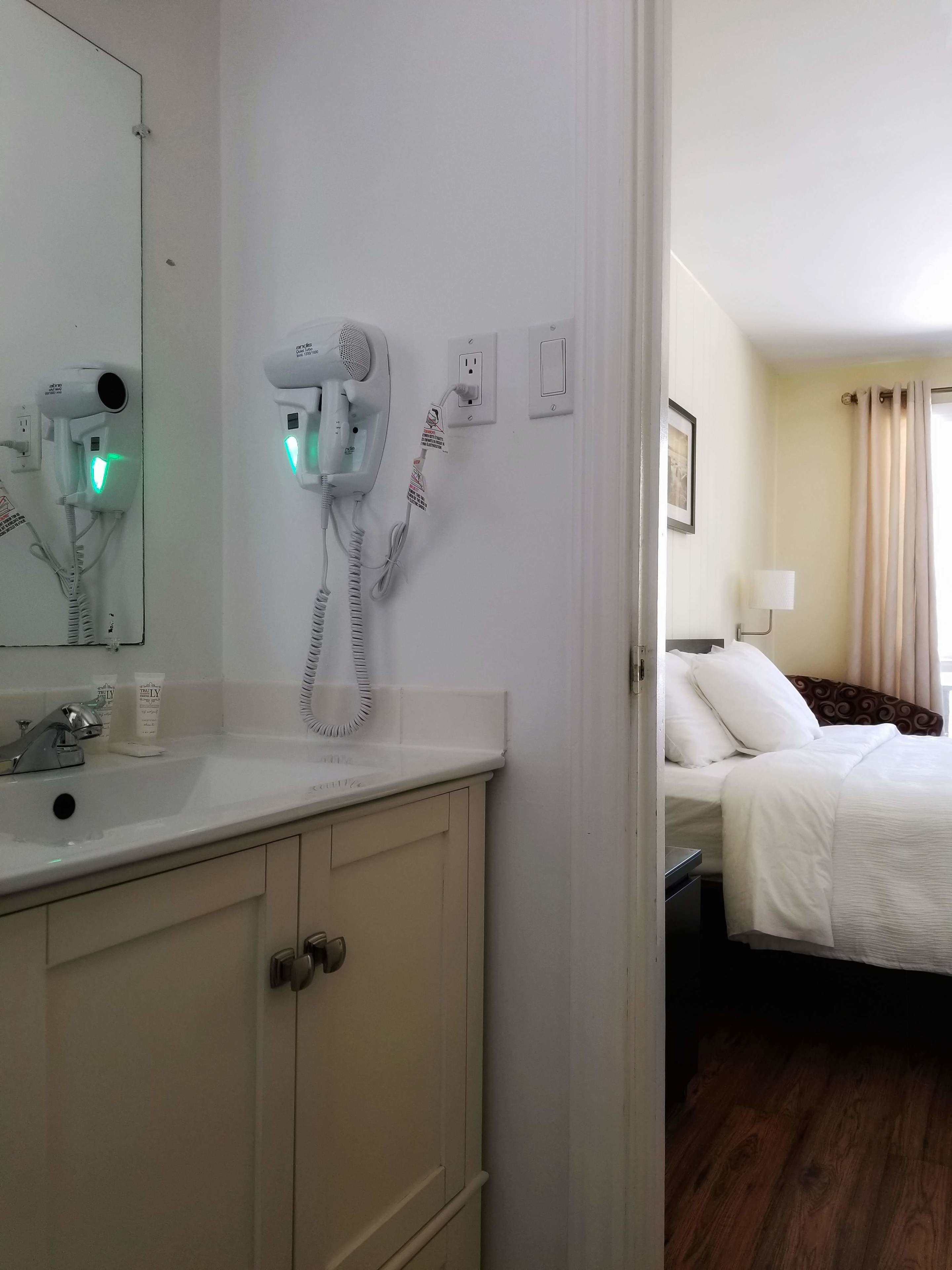 Ein Hotelbad mit Waschbecken, Spiegel, Fön und einem Bett im angrenzenden Zimmer.