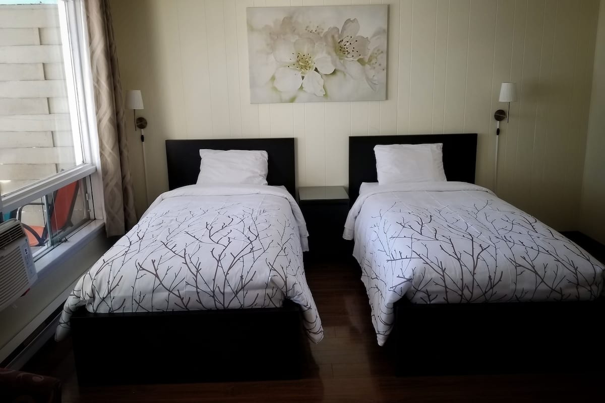 A hotel room with two beds, a window with blinds, a wall-mounted artwork, and a bedside table with a lamp.