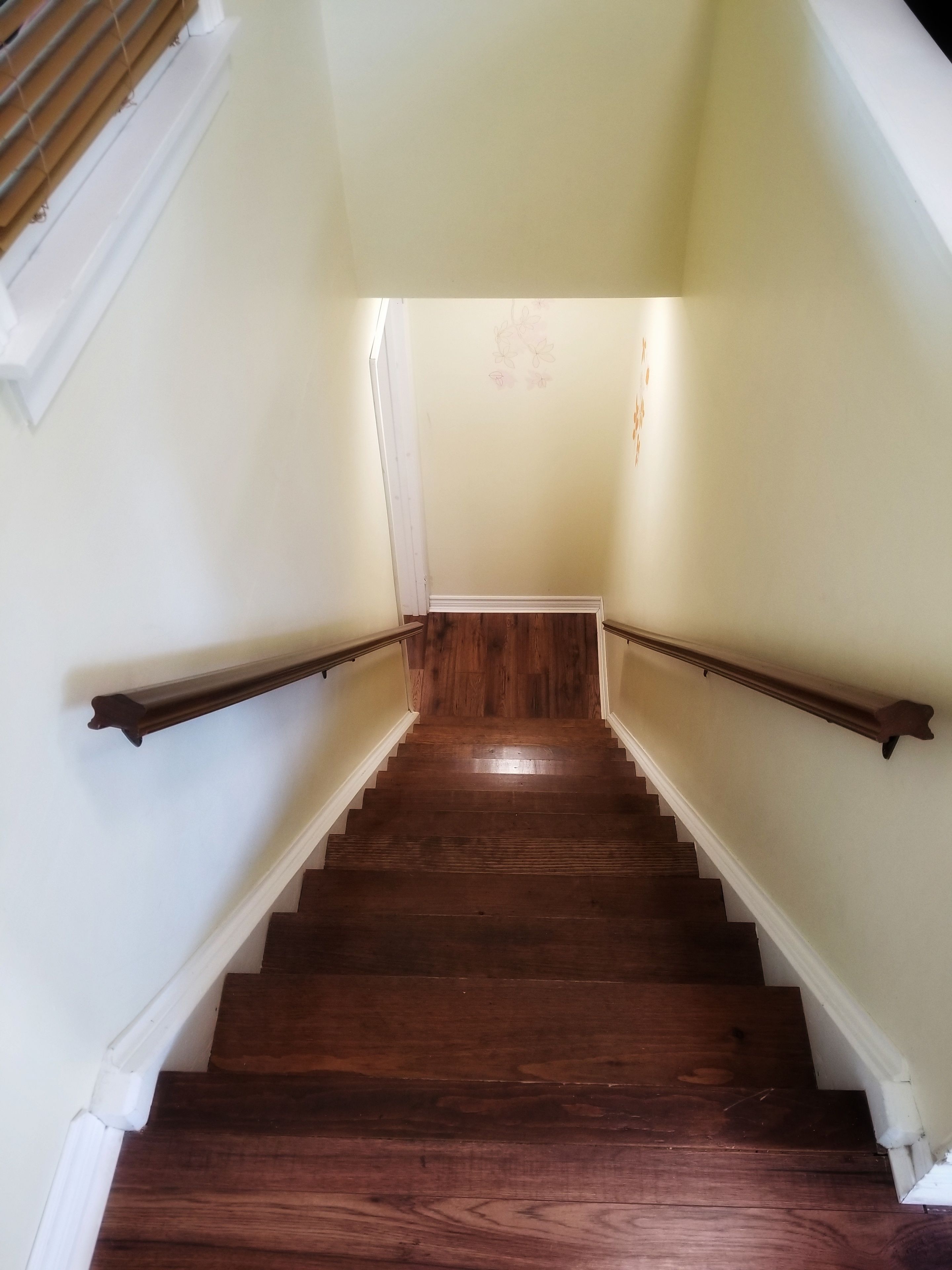Un escalier en bois, avec des moulures blanches et une rampe, mène à une pièce dont le mur est peint dans une couleur claire et orné d’un motif floral.