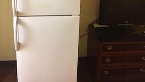 Un réfrigérateur blanc avec un four à micro-ondes posé sur le dessus, un téléviseur sur une commode en bois et un sol recouvert d’une moquette.