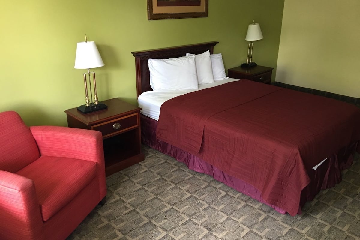 A hotel room with a large bed, two bedside tables with lamps, a red armchair, and a patterned carpet.