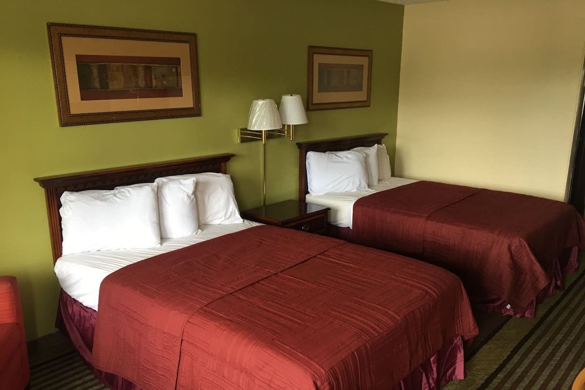 A hotel room with two beds, each with a wooden headboard and red bedding. There are two bedside tables with lamps, and the walls are painted green.