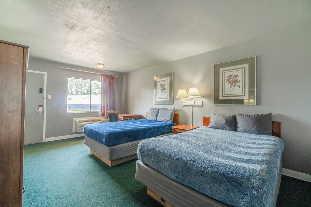 Habitación de hotel con dos camas, un escritorio, una silla, un ventanal con cortinas y cuadros en la pared.