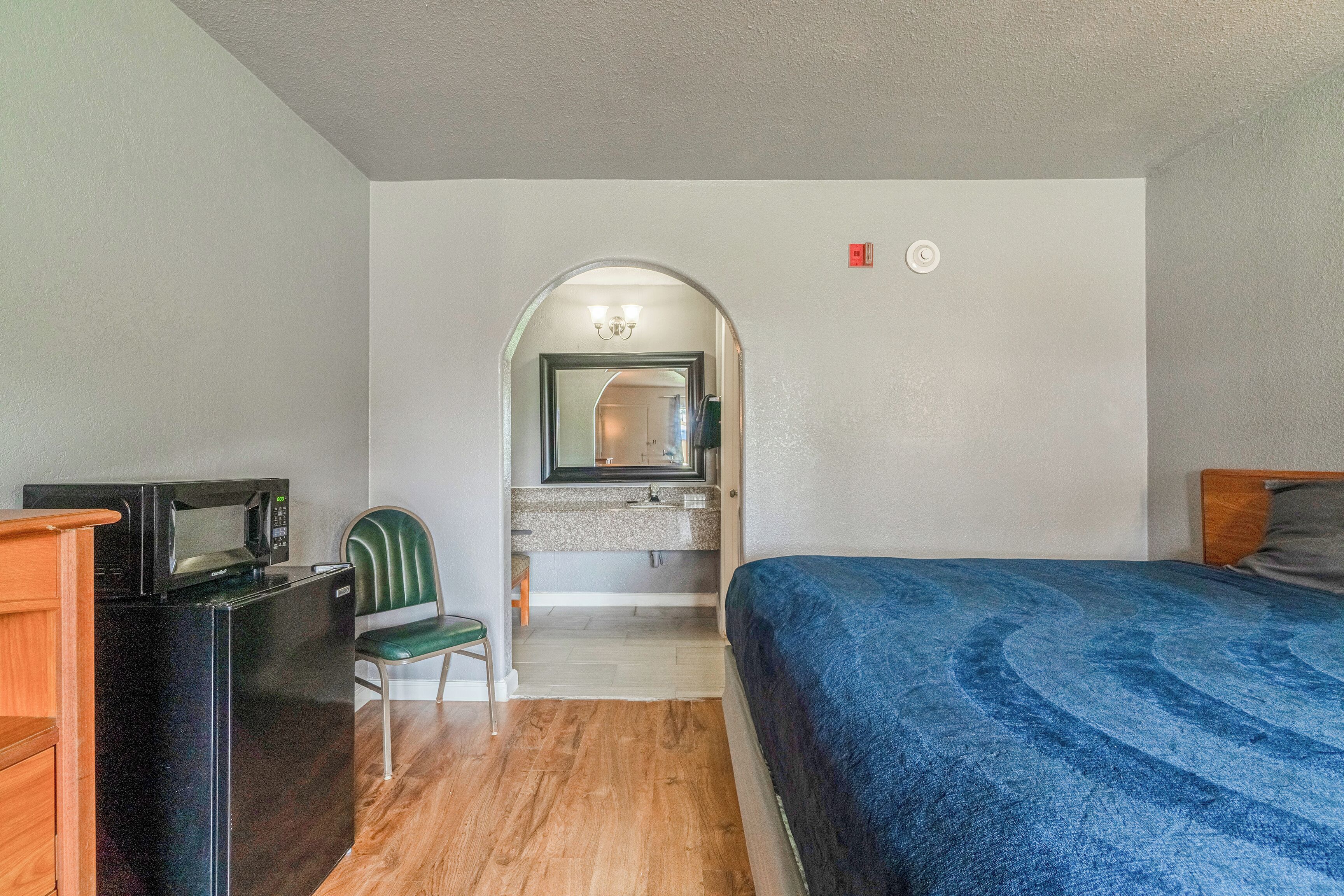 Une chambre d’hôtel compacte comprenant un lit, un bureau avec un micro-ondes, une chaise, une petite salle de bain avec un lavabo et un miroir, et une tête de lit en bois.