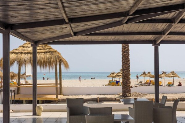 Un espacio al aire libre con mesas y sillas, con vista a una playa con sombrillas y gente.