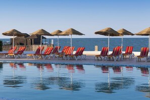 Área da piscina com espreguiçadeiras e guarda-sóis de palha, uma estrutura de madeira e vista para o mar.