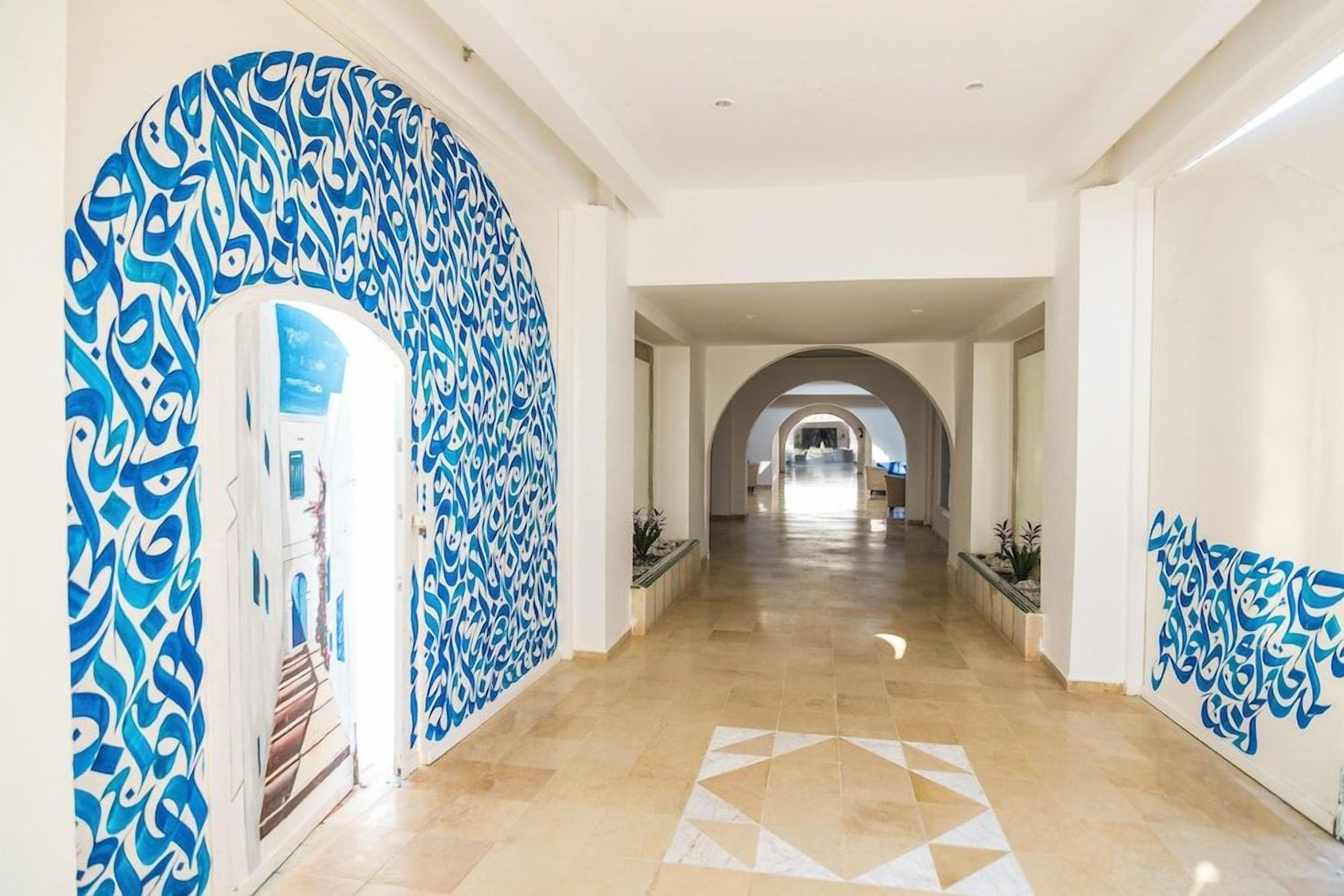 Un couloir d’hôtel avec un sol à motifs géométriques, des murs blancs et des éléments décoratifs bleus.