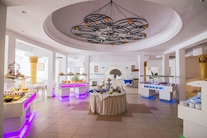 Um amplo hall de hotel com uma mesa redonda central, várias estações de buffet e um lustre de teto marcante.