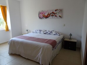 A bedroom with a bed, a nightstand, a lamp, and a wall art piece. - La Salamandra (Puerto Aventuras)