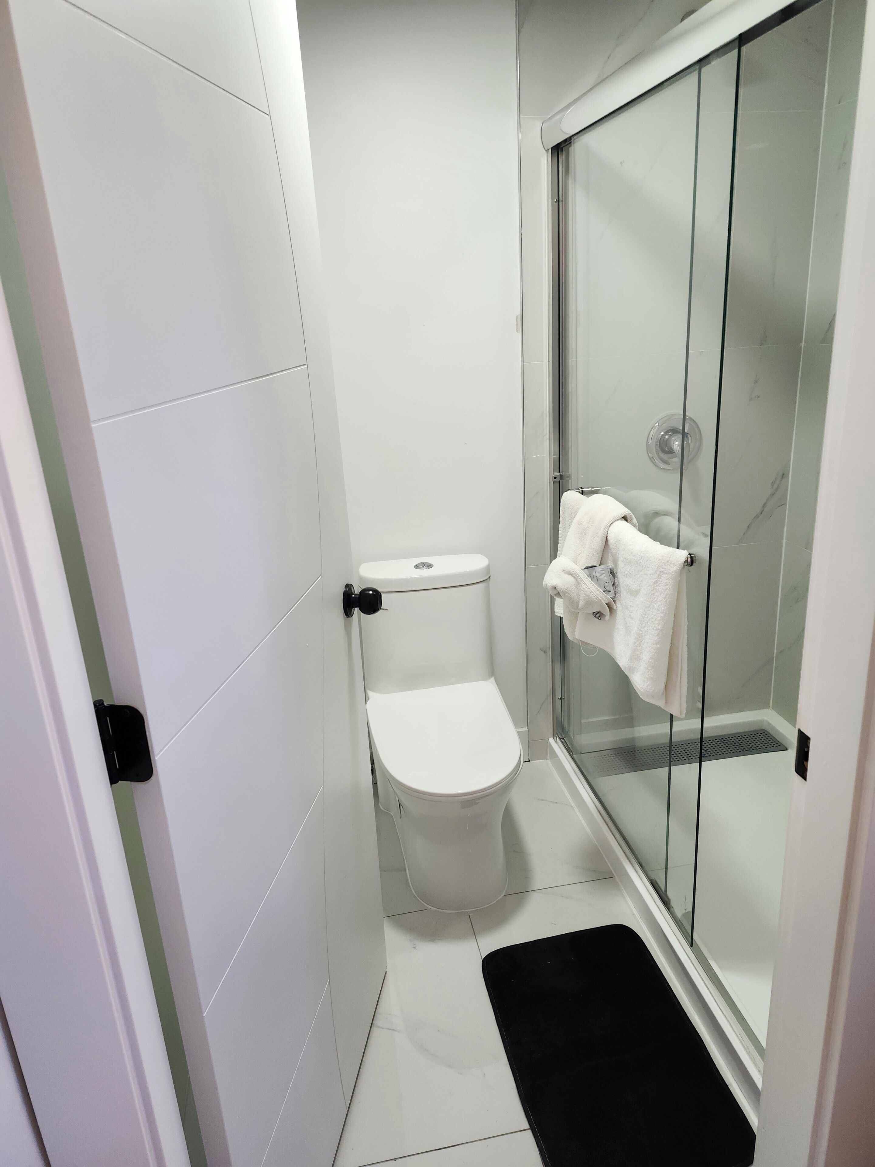 A small, modern bathroom with a toilet, glass shower enclosure, and a black mat.