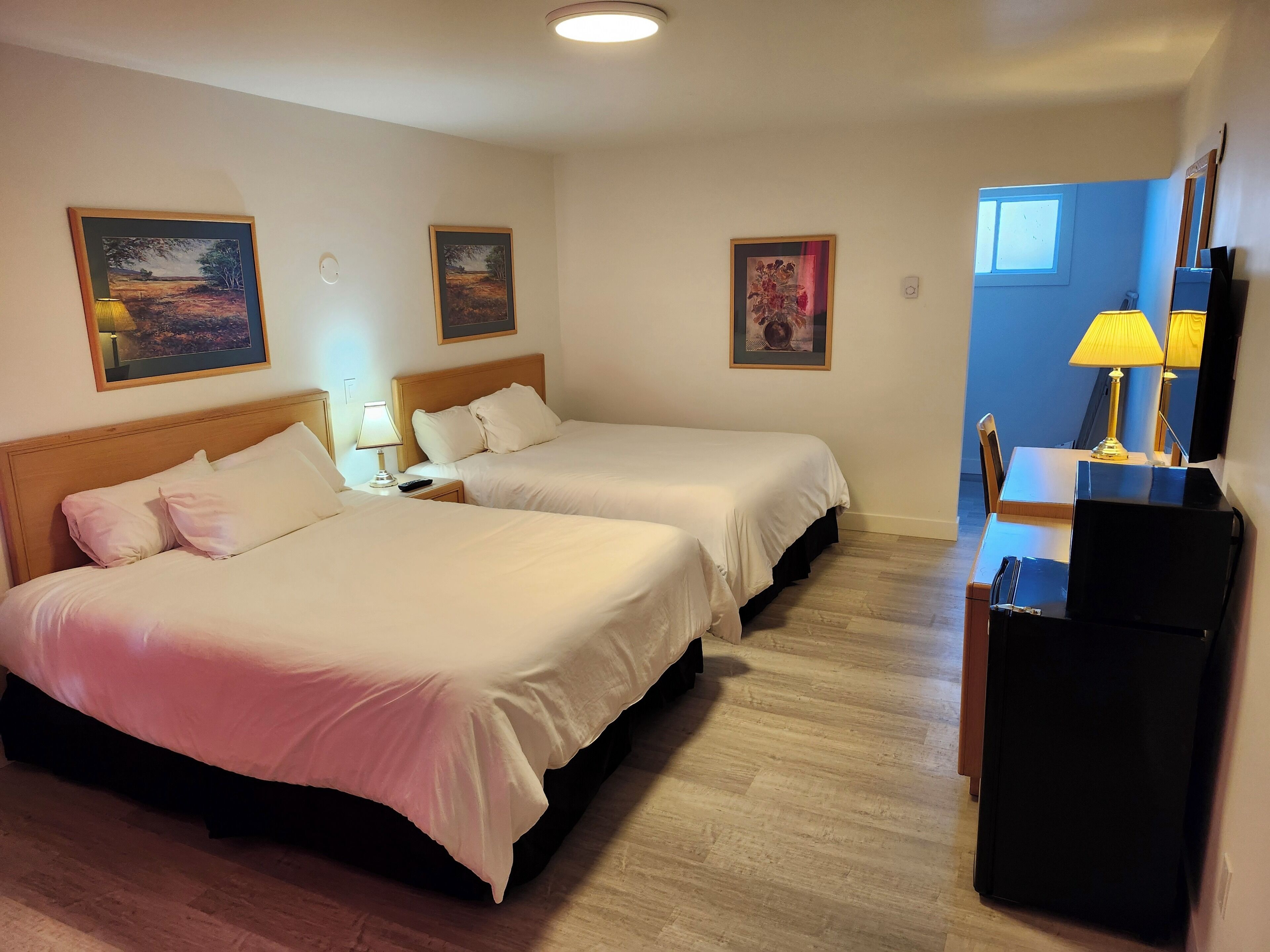 A hotel room with two beds, a desk, a chair, a lamp, and a TV.