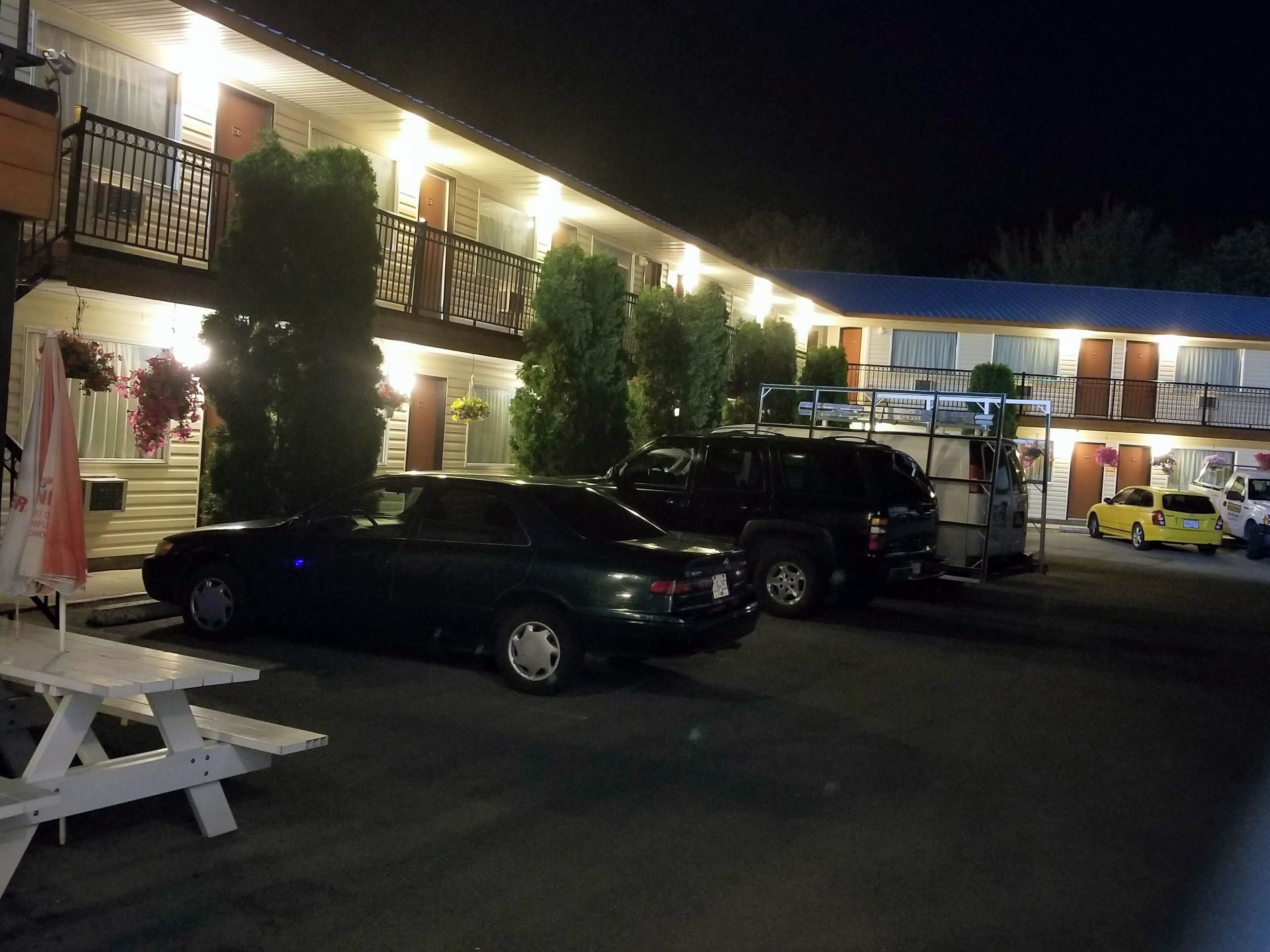 Un motel con área de estacionamiento techada, una mesa de picnic y varios vehículos estacionados al frente.