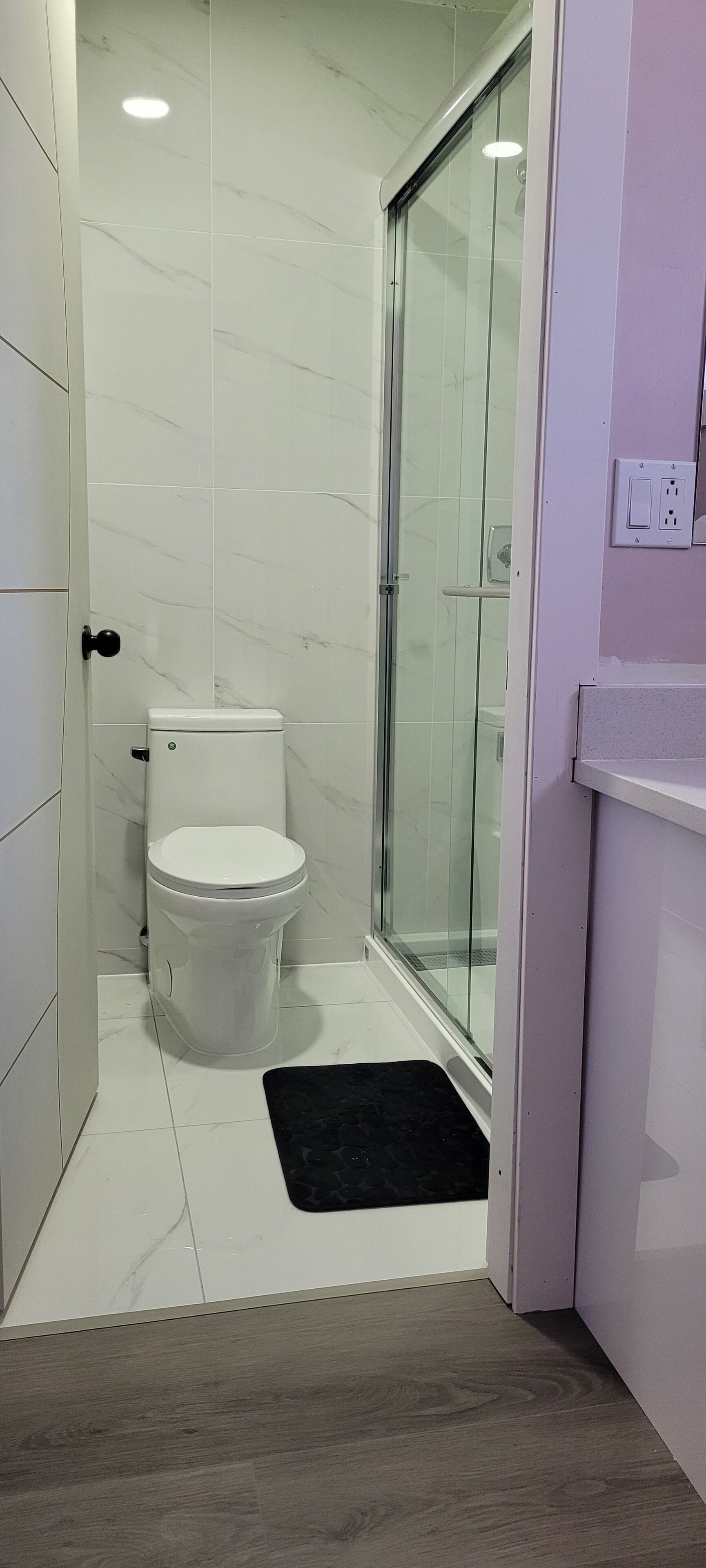 A bathroom with a toilet, a glass shower enclosure, and a black mat on the floor.
