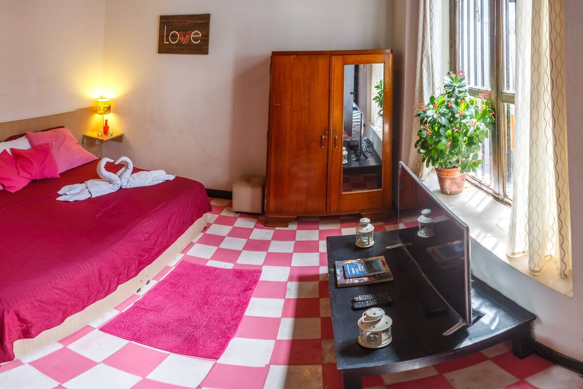 A hotel room with a checkered floor, a bed with pink bedding, a wooden wardrobe, a small table with a lamp, and a potted plant by the window.
